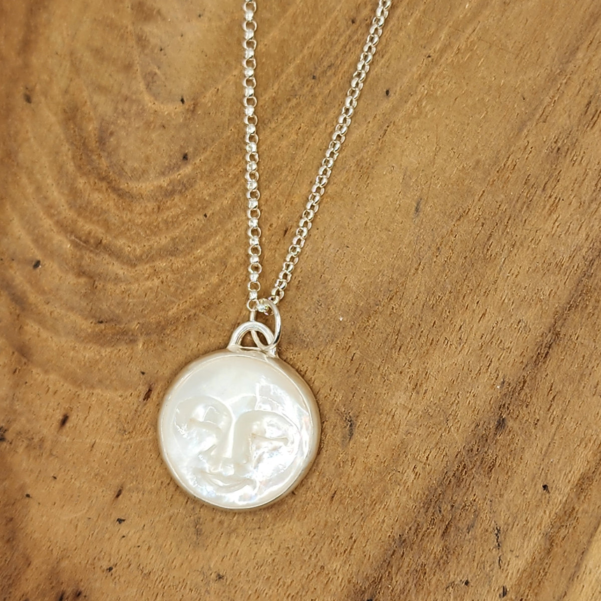 A sterling silver necklace with a round, mother of pearl with carved moon face, displayed on a natural wood background.