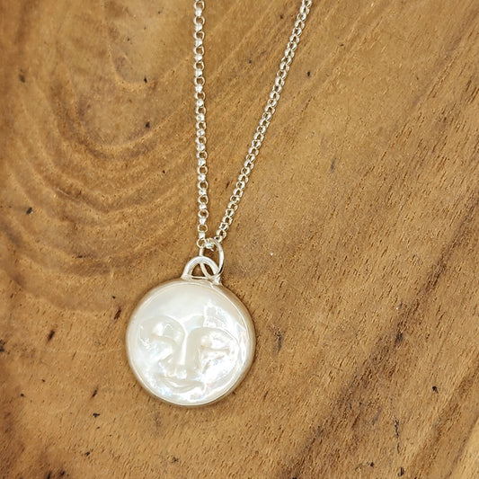 A sterling silver necklace with a round, mother of pearl with carved moon face, displayed on a natural wood background.