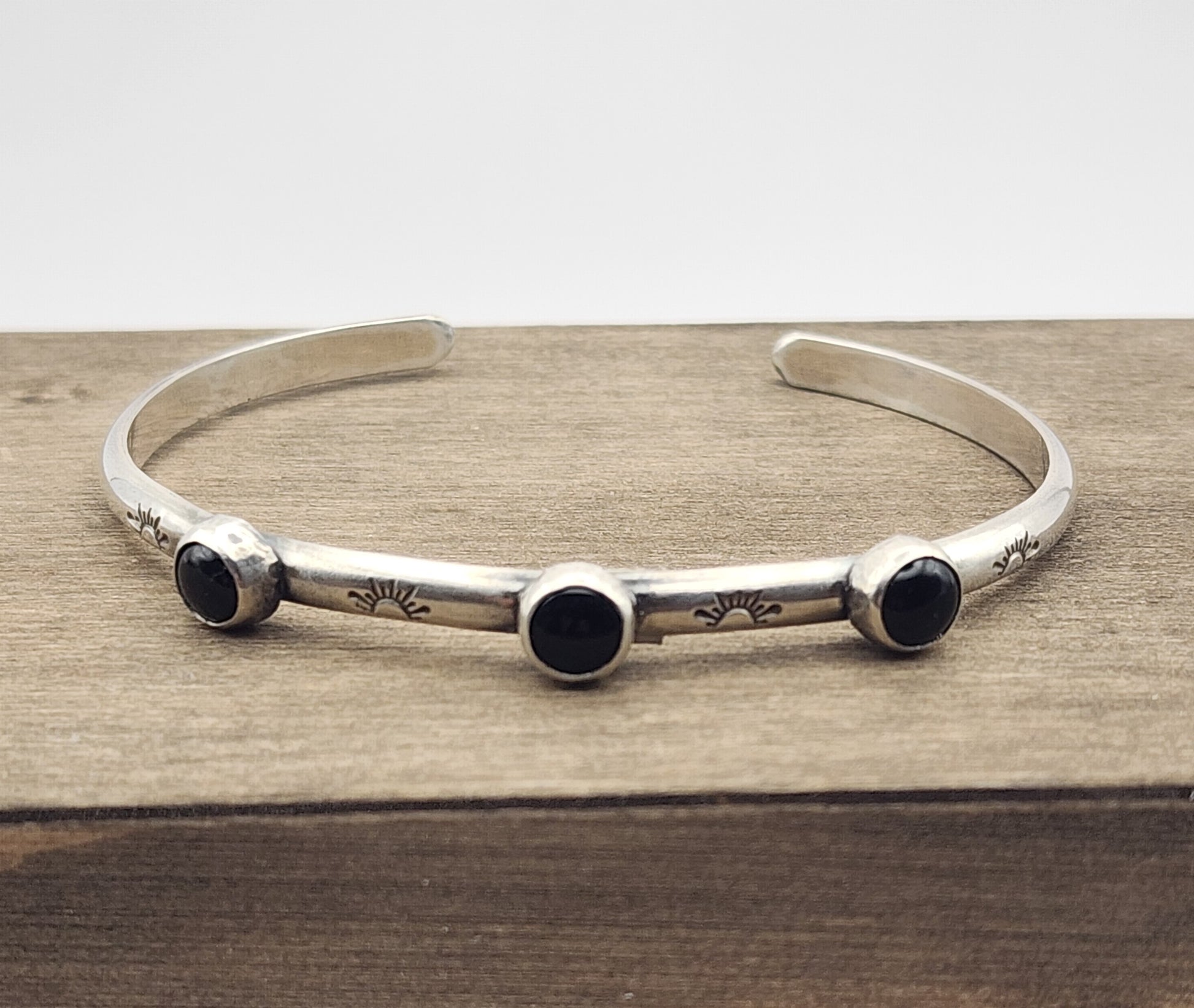 Sterling silver cuff bracelet with four stamped half suns with rays with three small onyx stones between the stamps on a wooden block and a white background
