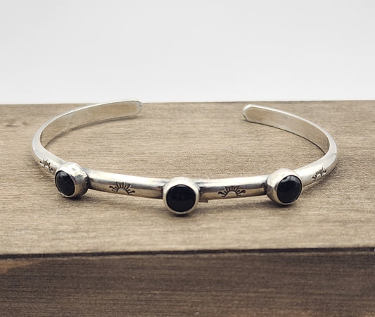 Sterling silver cuff bracelet with four stamped half suns with rays with three small onyx stones between the stamps on a wooden block and a white background