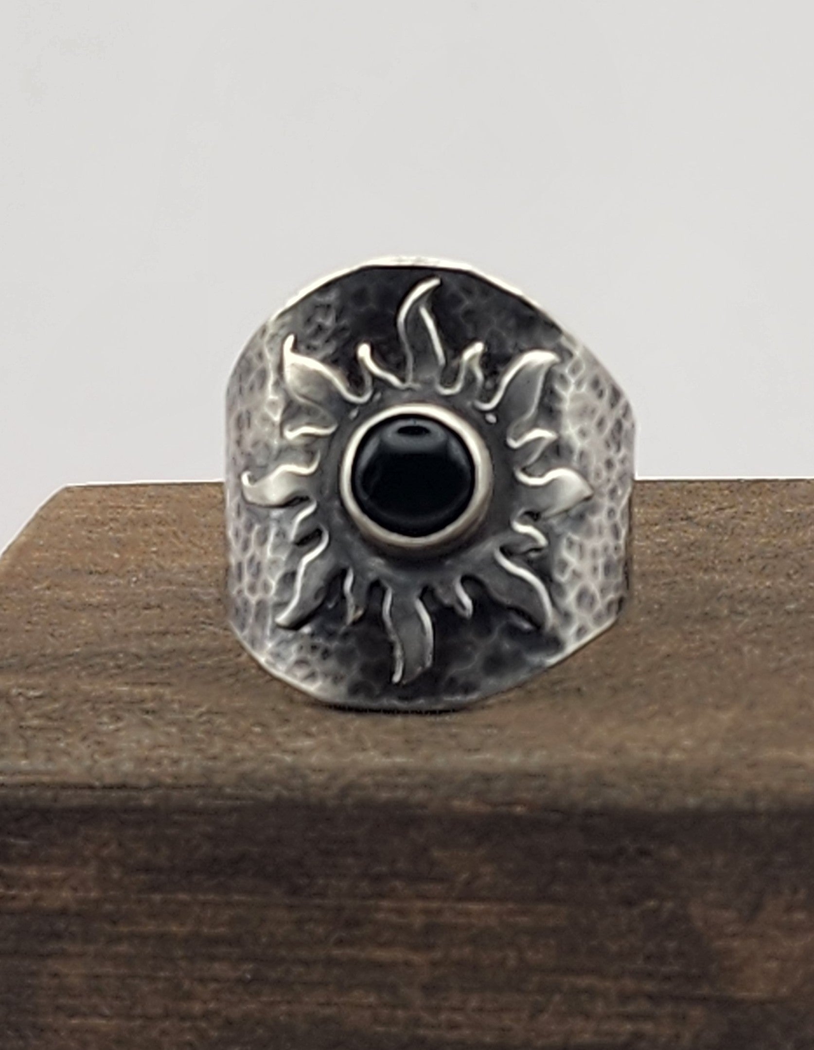 A hammered silver ring featuring a central black onyx stone surrounded by a sunburst motif, displayed on a dark wooden block against a white background.
