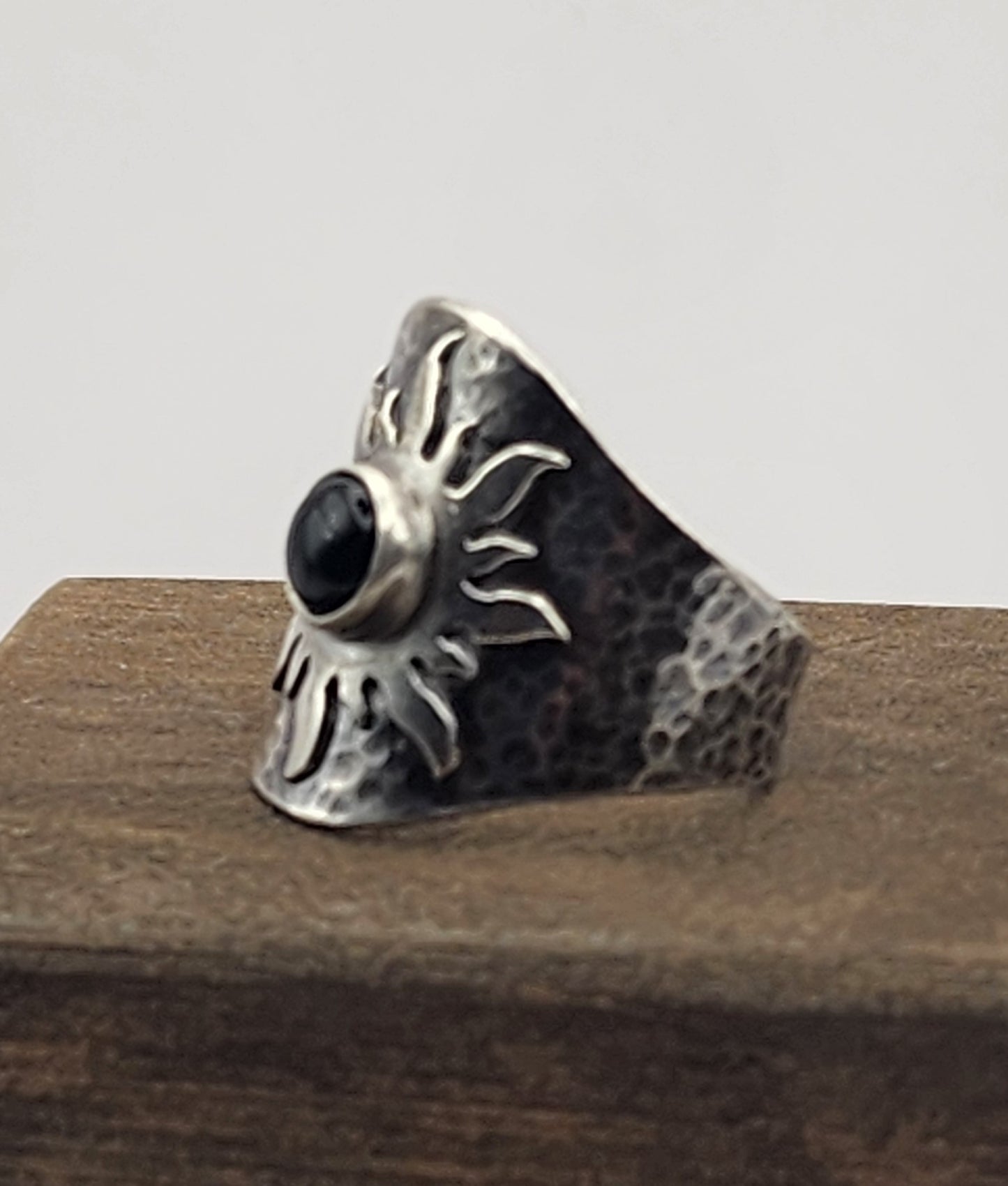 A hammered sterling silver ring with a central black onyx stone, displayed on a dark wooden block against a white background. Side view of ring.