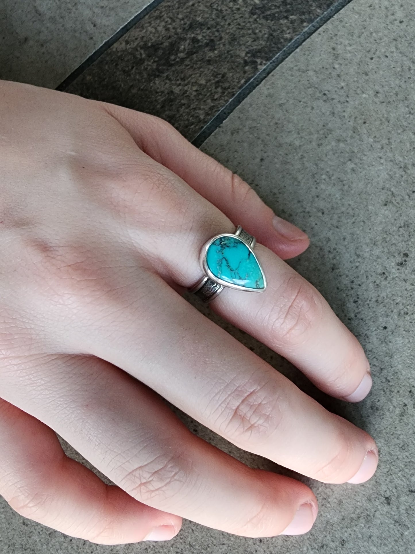 Hand wearing a teardrop shaped turquoise silver ring on a concrete surface