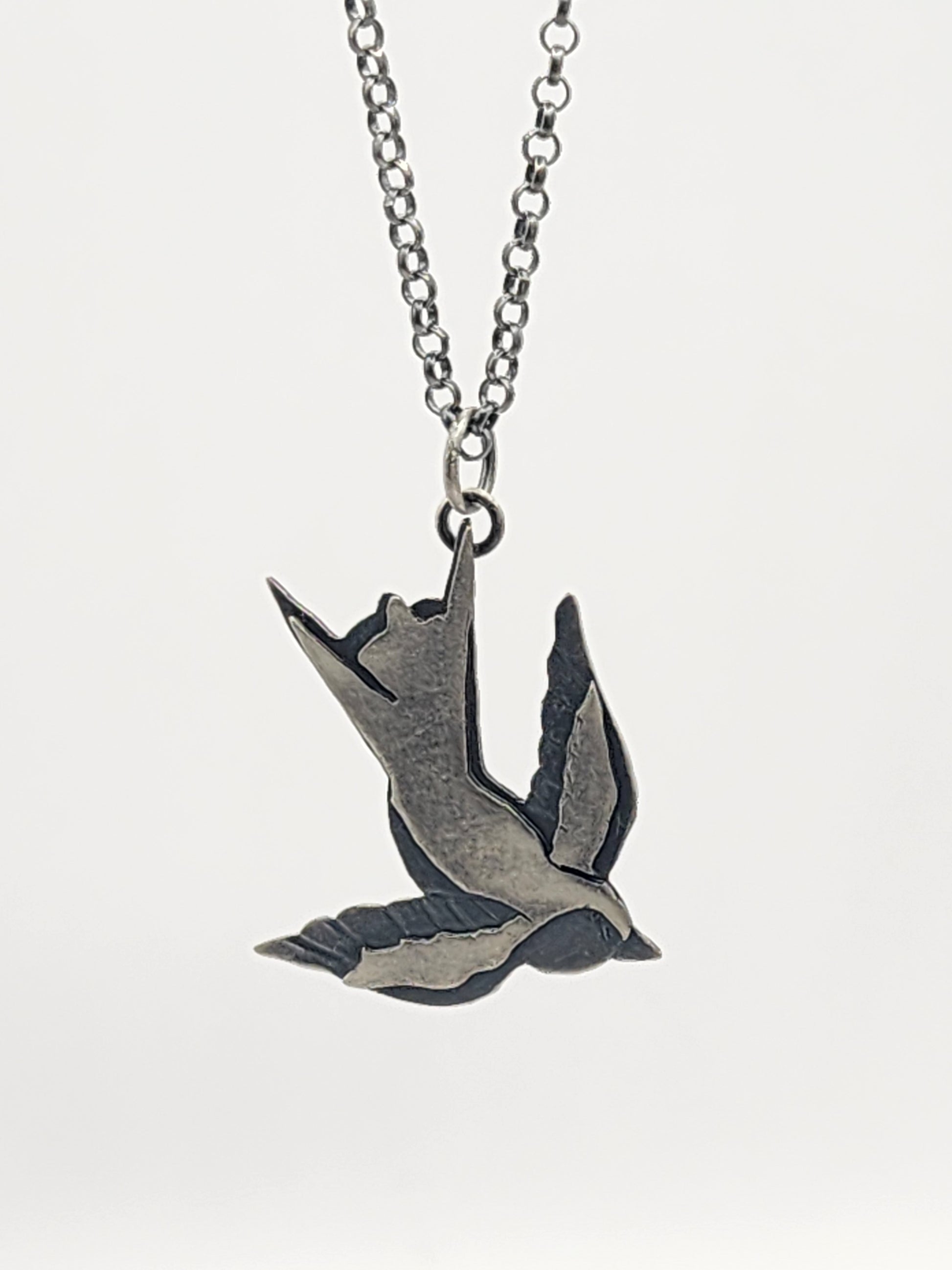 A silver-toned metal pendant necklace featuring a flying bird design, suspended from a matching chain against a white background.