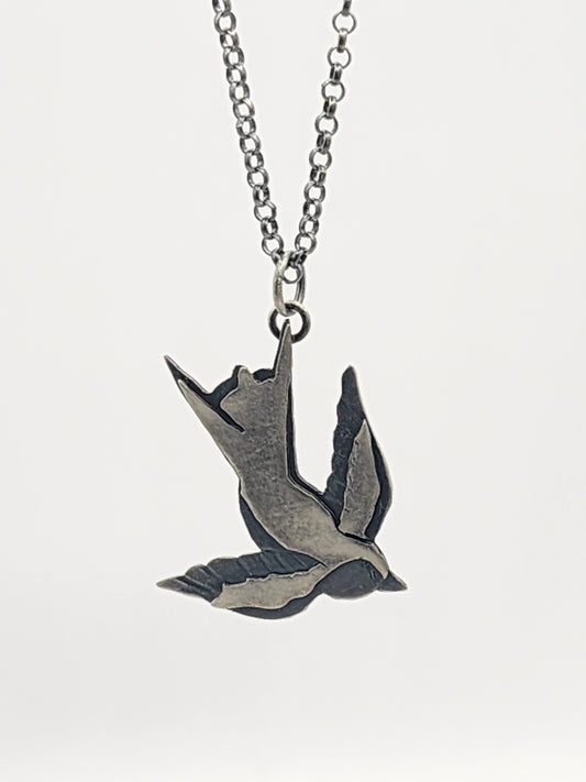 A silver-toned metal pendant necklace featuring a flying bird design, suspended from a matching chain against a white background.