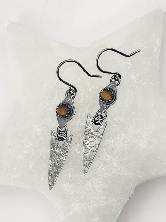 A pair of handmade dangle earrings with hammered silver arrowhead/chevron charms, small tigers eye stones, and french earwires, displayed on a textured white surface.