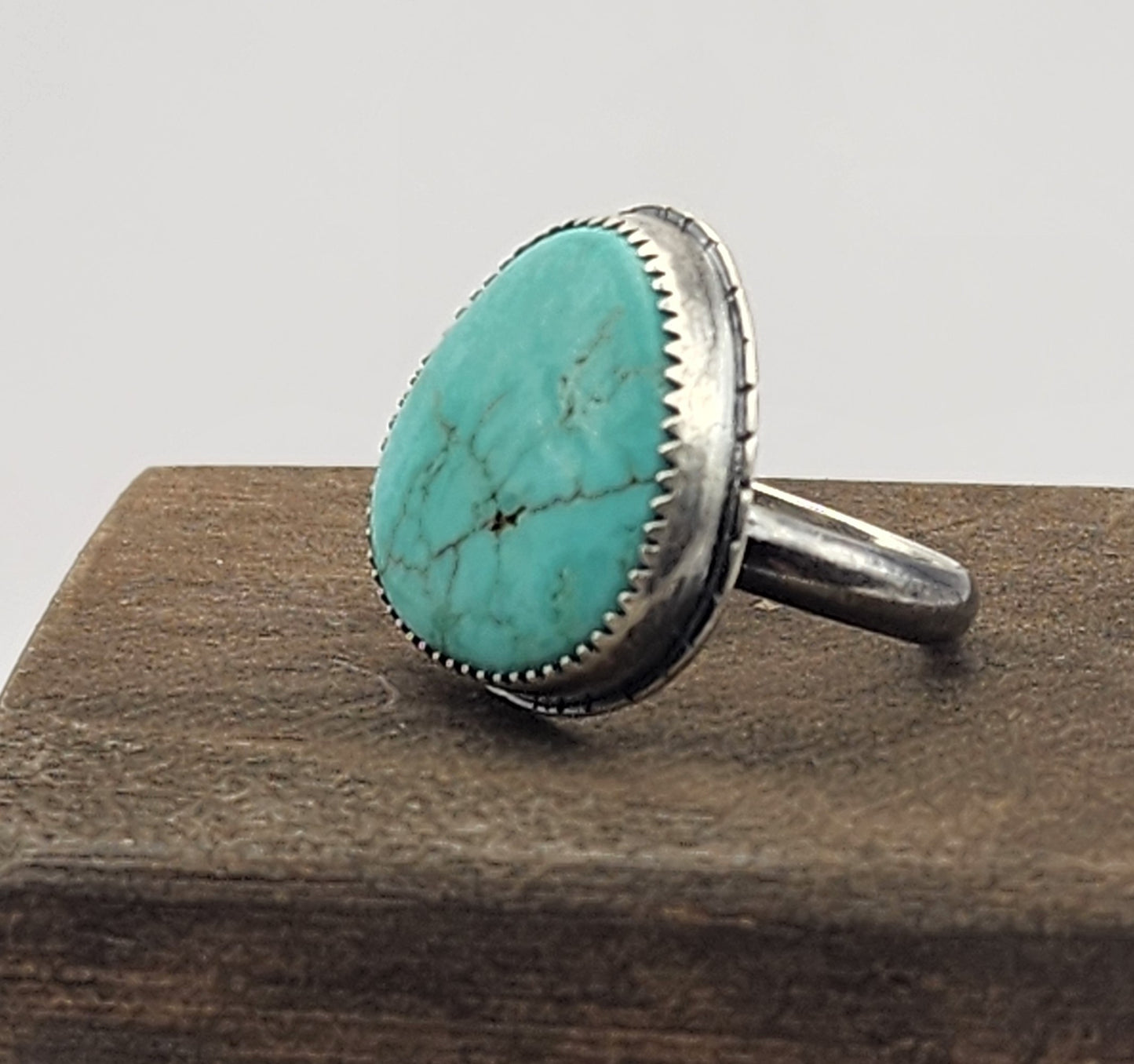 A sterling silver ring featuring a large, oval/teardrop-shaped turquoise stone with natural matrix veining, displayed on a dark wood block against a white gray background.