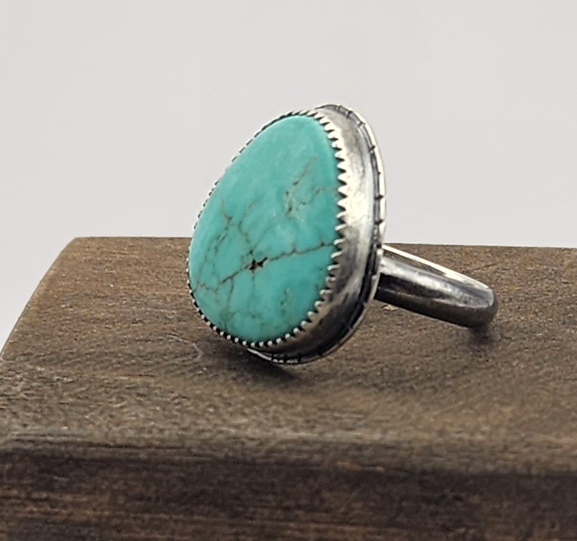 A sterling silver ring featuring a large, oval/teardrop-shaped turquoise stone with natural matrix veining, displayed on a dark wood block against a white gray background.