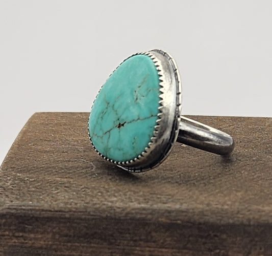 A sterling silver ring featuring a large, oval/teardrop-shaped turquoise stone with natural matrix veining, displayed on a dark wood block against a white gray background.