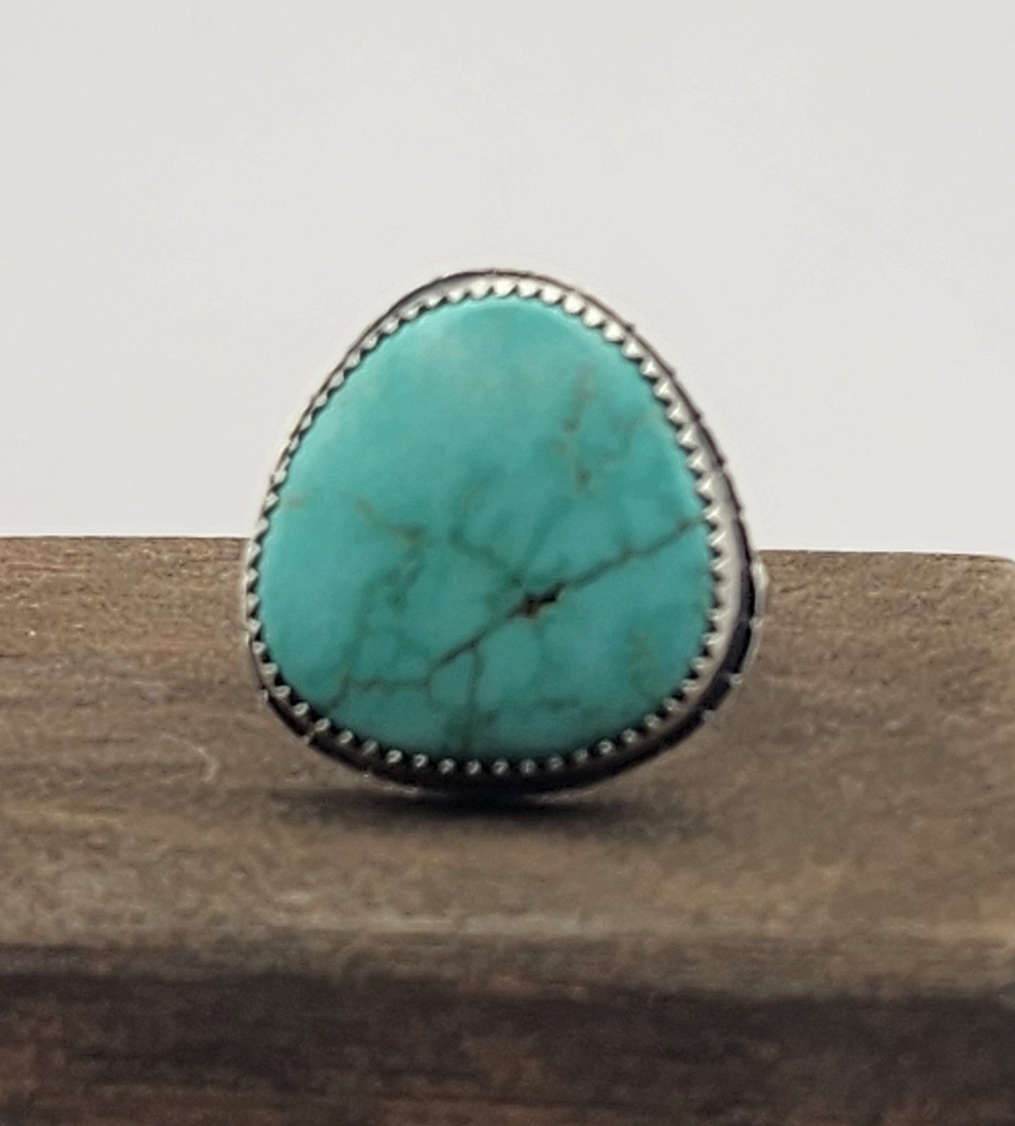 A sterling silver ring featuring a large, oval/teardrop-shaped turquoise stone with natural matrix veining, displayed on a textured brown surface against a white background.