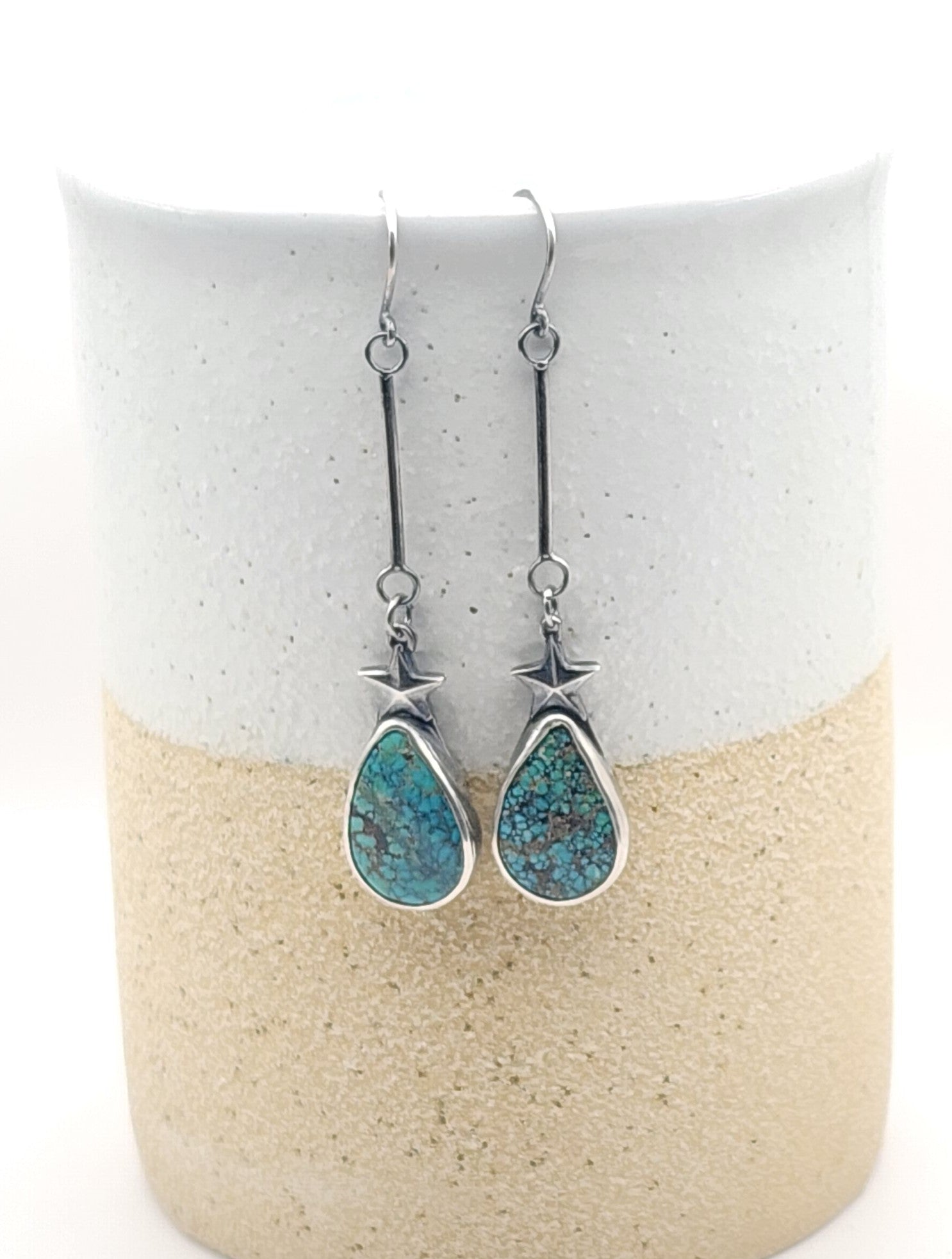 A pair of silver dangle earrings with teardrop-shaped turquoise-blue stone pendants and star-shaped silver accents, displayed on a two-toned ceramic stand against a white background.